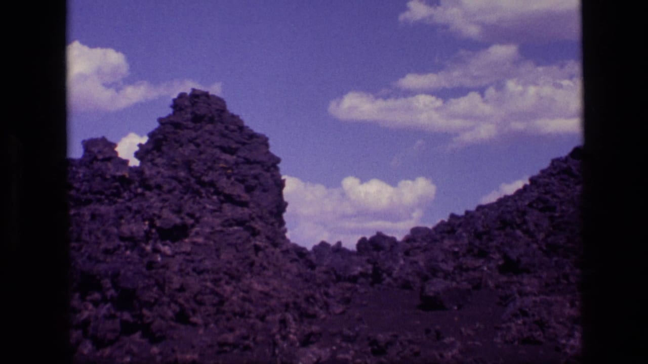 1972 Idaho 8mm view of a tall, rough volcanic formation and uneven lava terrain below a bright clouded sky.
