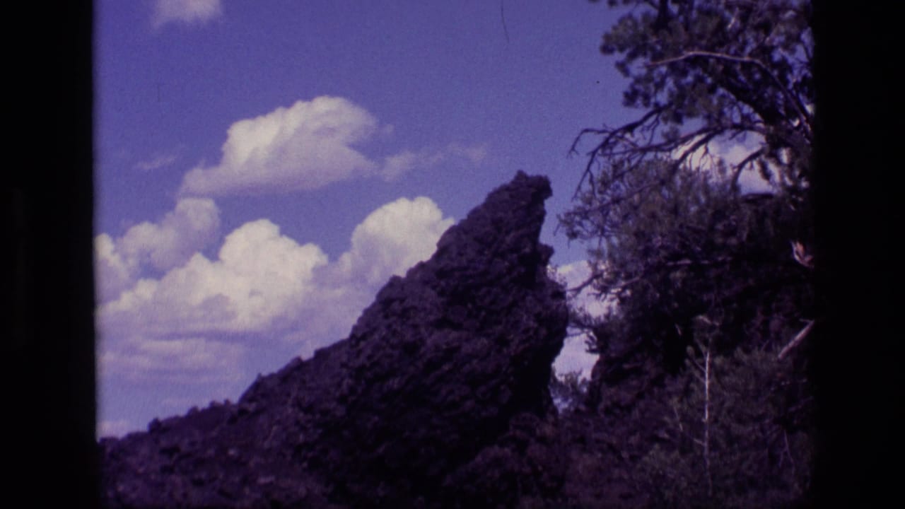 1972 Idaho 8mm frame showing a jagged rock outcrop, dark vegetation, tree branches, and bright clouds in a saturated sky.