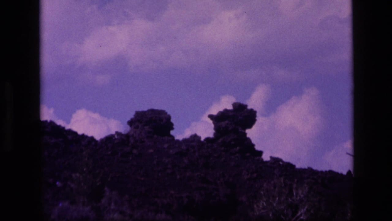 1972 Idaho 8mm frame of a shadowed, rugged hillside against a purple-blue sky with soft cloud forms and heavy film grain.