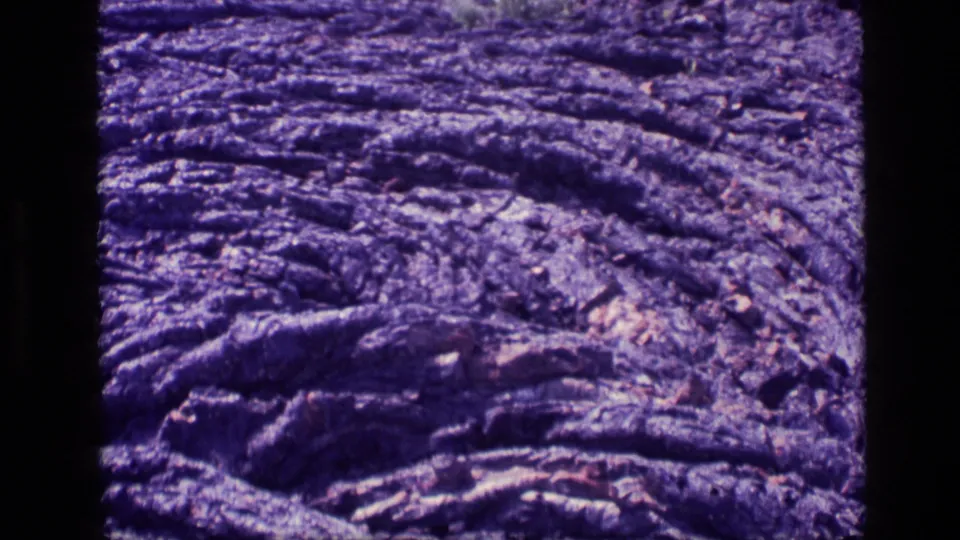 Close-up 8mm view of ridged cooled lava in Idaho, with dense grain, magenta-purple cast, and no visible people.