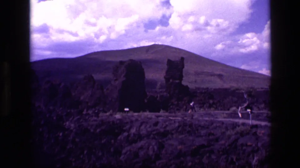 Wide 1972 Idaho home-movie view of dark lava formations, sparse ground, small signs, and a distant mountain beneath clouds.