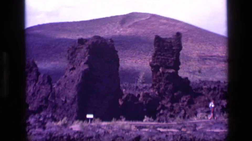1972 Idaho 8mm frame showing tall dark lava formations, a rounded mountain behind them, signs, and small visitors at the site.
