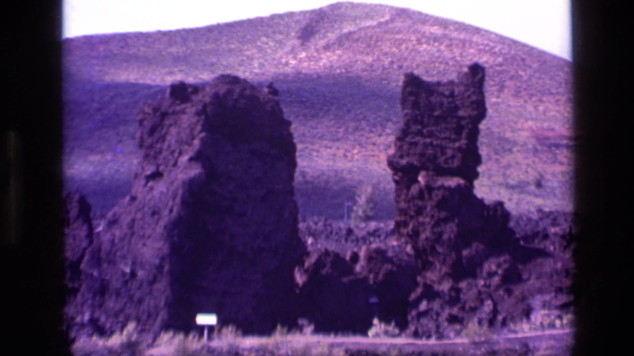 1972 Idaho 8mm frame showing dark volcanic-looking rock spires, low scrub, a small sign, and distant mountain slopes.