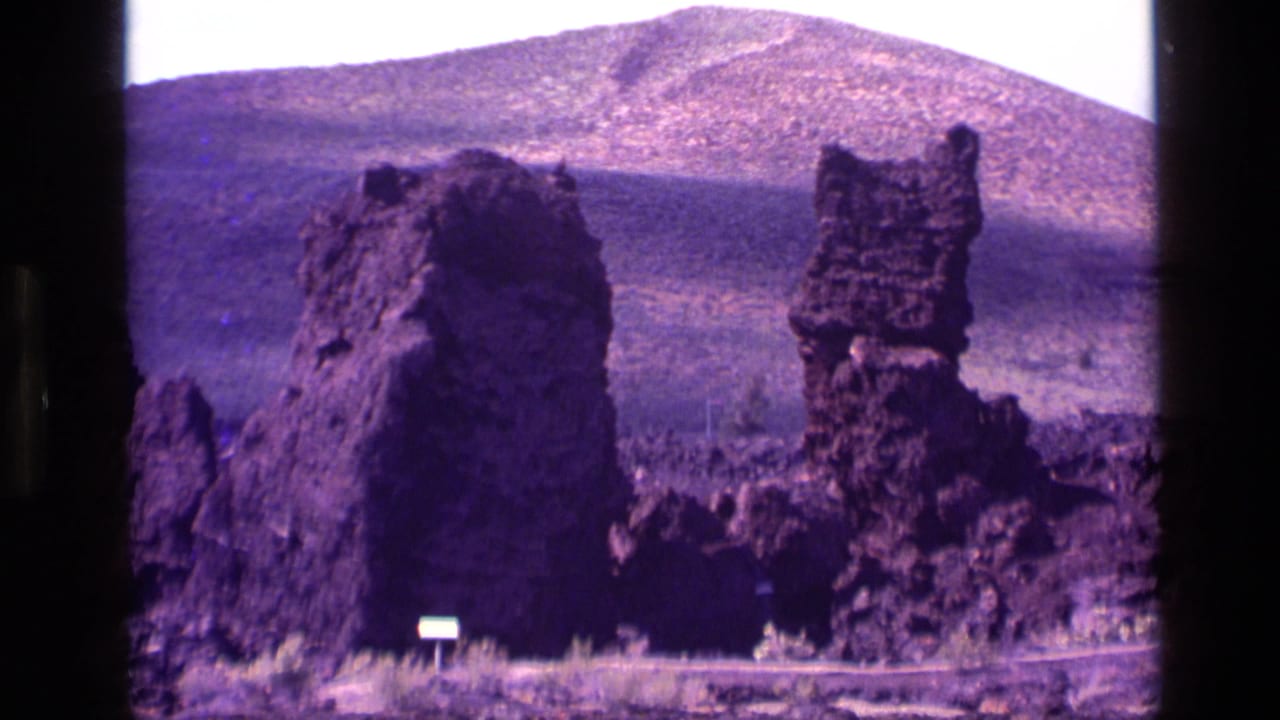 1972 Idaho home-movie frame with jagged dark rocks, a broad mountain backdrop, pale sky, and visible 8mm frame edges.