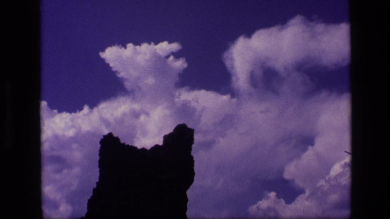 1972 Idaho 8mm frame showing a dark jagged rock formation silhouetted below puffy clouds in a saturated purple-blue sky.
