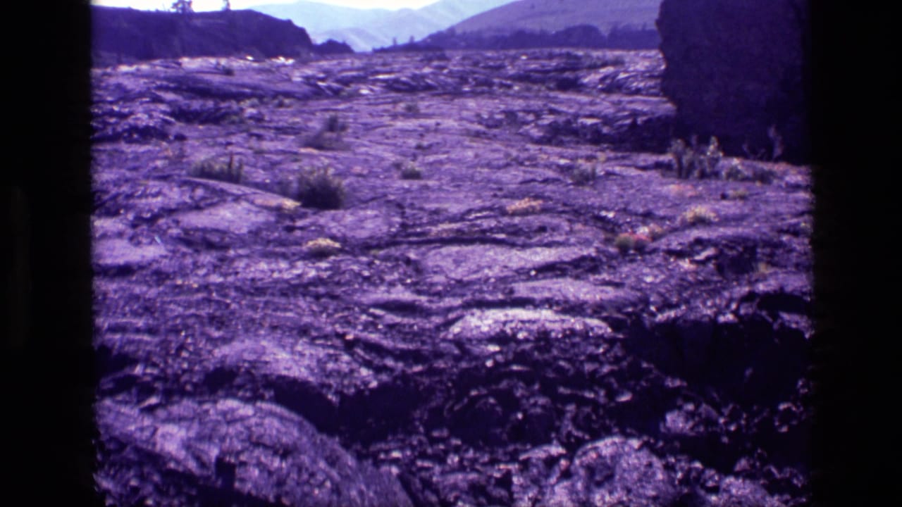 1972 Idaho 8mm frame showing dark rocky terrain, sparse vegetation and distant mountain shapes with a purple film cast.