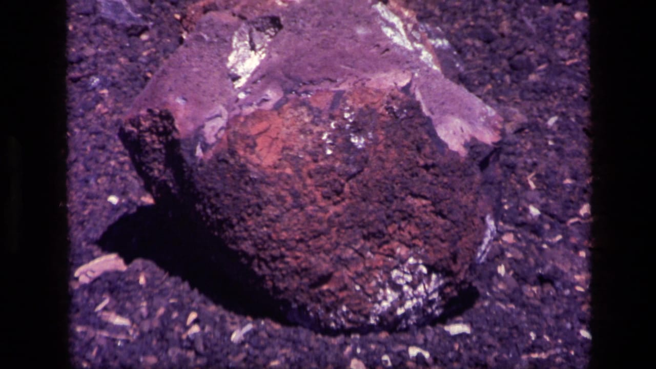 Close view of dark reddish rock with shiny flecks on gravelly ground.