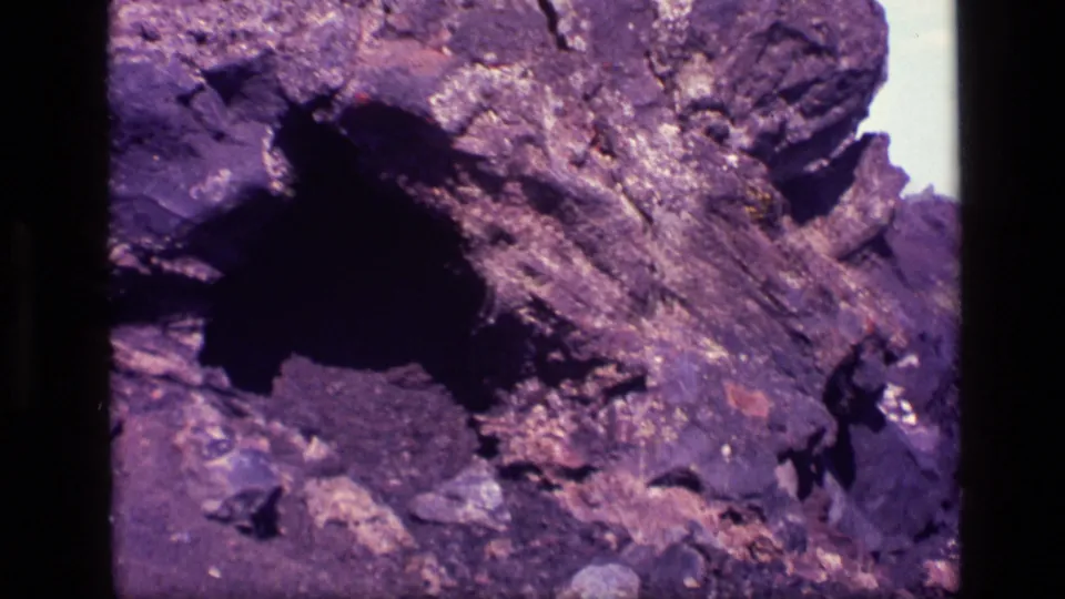 1972 Idaho frame showing a rough black and brown rock outcrop with a small cave-like hollow in deep shadow.