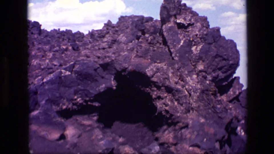 8mm Idaho landscape frame showing dark volcanic-looking rocks, a small shadowed hollow, and pale sky at the edge of the outcrop.