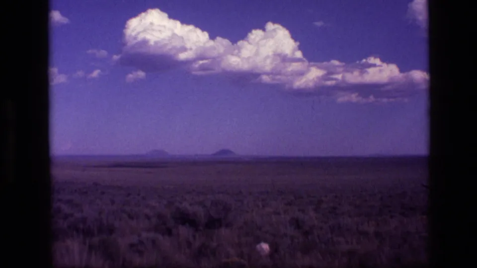 1972 Idaho home-movie frame with low brushland, distant mountain silhouettes, and bright clouds under a purple-tinted sky.