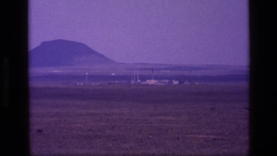 1972 Idaho 8mm frame with a distant mountain, open dry plain, and small far-off structures near the horizon.