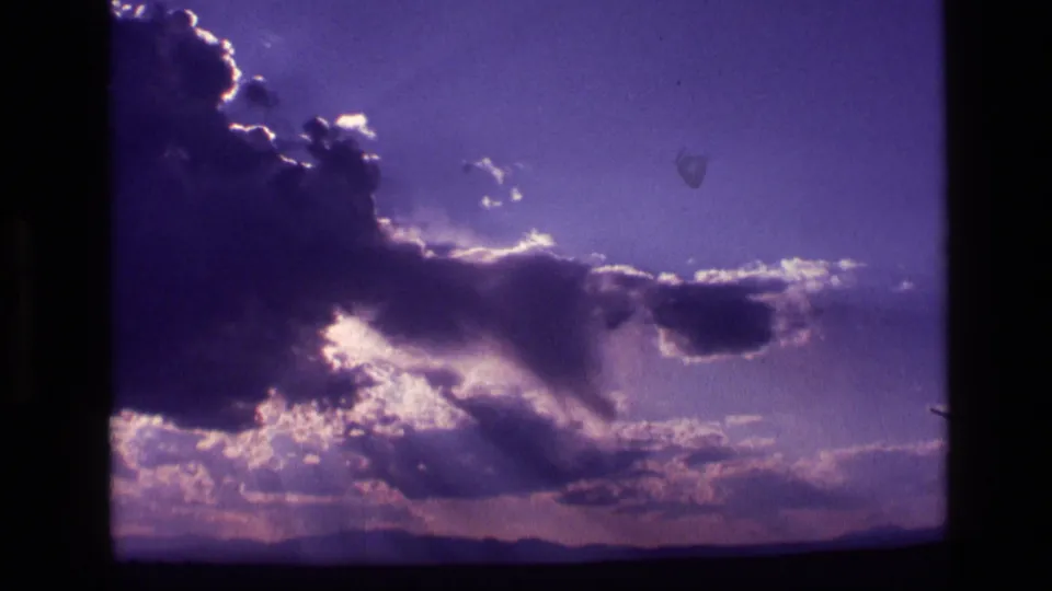 1972 Idaho 8mm frame showing dark clouds, bright sunbeams, and a low landscape horizon in purple-shifted color.