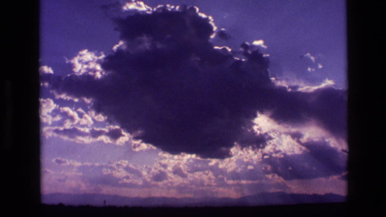 1972 Idaho 8mm frame with a voluminous backlit cloud, scattered smaller clouds, sun rays, and a low mountain horizon.