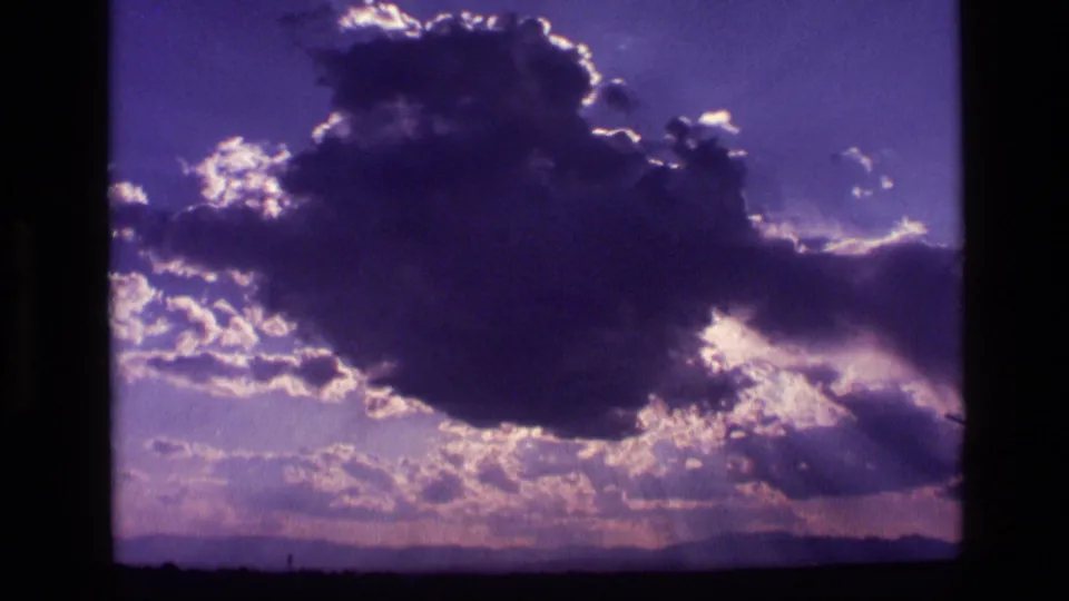 1972 Idaho 8mm frame with a voluminous backlit cloud, scattered smaller clouds, sun rays, and a low mountain horizon.