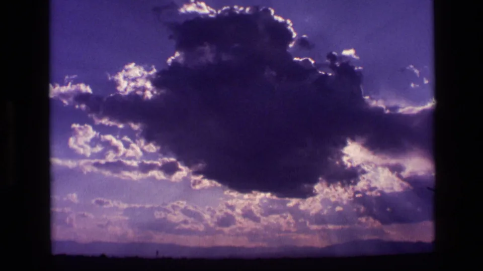 1972 Idaho 8mm frame showing a broad cloud formation with bright rim light, soft haze, purple color shift, and a low horizon.