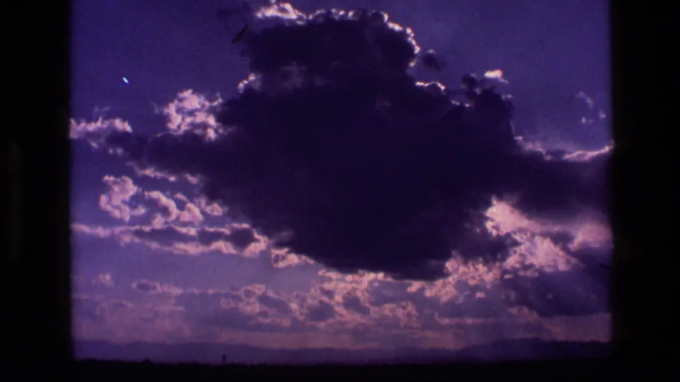 1972 Idaho 8mm frame with a dark cloud silhouette, pink-purple sky tones, bright breaks of light, and a low distant ridge.