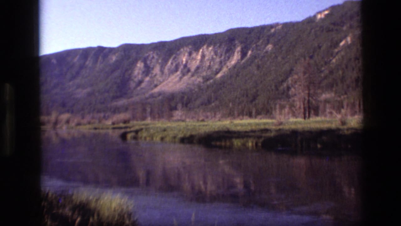 1972 Idaho 8mm view of a forested mountain ridge, grassy shoreline, and still water framed by dark film edges.