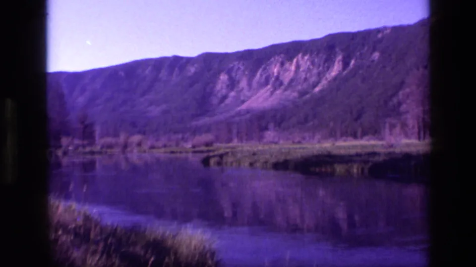 Purple-shifted 1972 Idaho 8mm frame showing a mountain ridge, low shoreline vegetation, and water reflections.