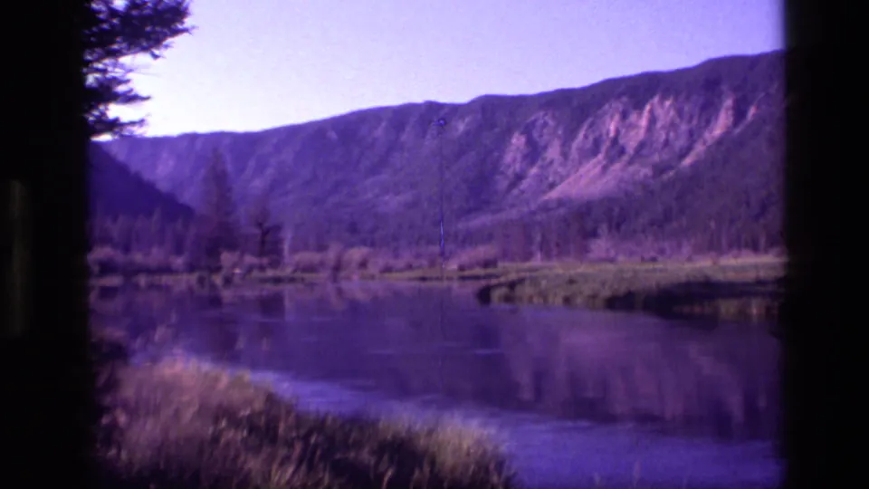 1972 Idaho 8mm frame of calm water below a forested mountain ridge, with a nearby tree edge and dark film border.