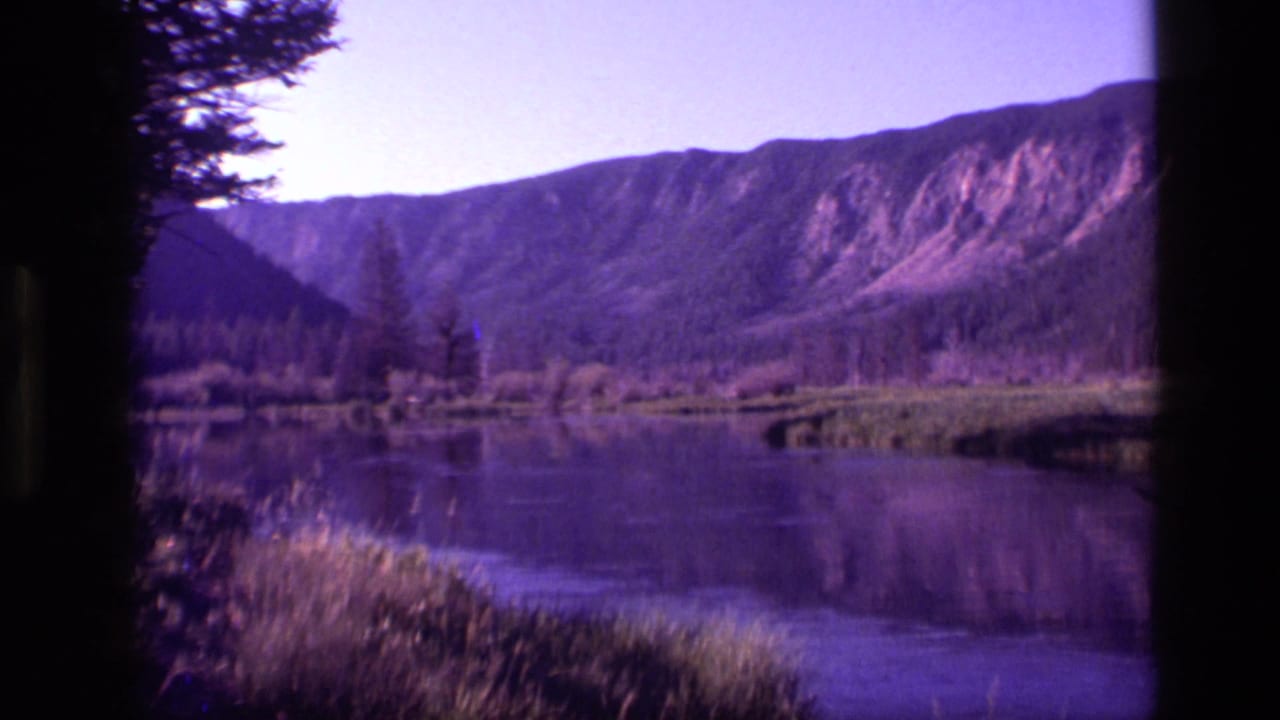 Calm lake below a mountain ridge with trees and shoreline grass, framed by dark film edges.