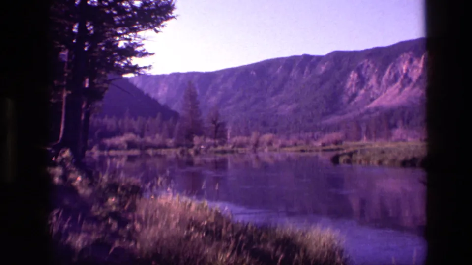 1972 Idaho 8mm landscape frame with foreground grass, reflective lake water, trees, and distant mountains.