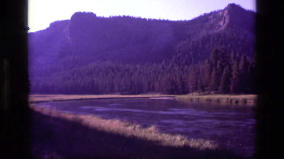 1972 Idaho 8mm frame showing a broad lake or wetland foreground below pine forest and a rugged mountain ridge.