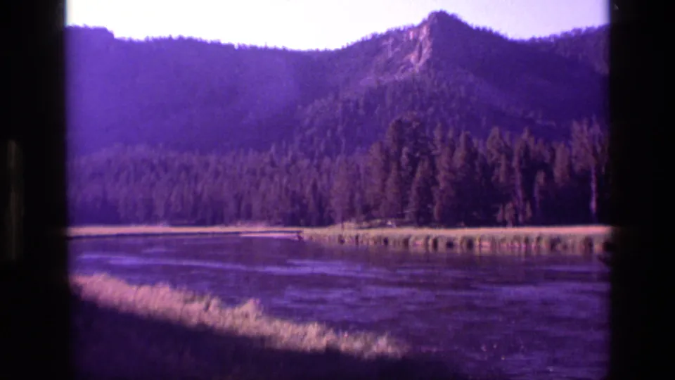 1972 Idaho 8mm frame showing a pointed mountain rise, dense pine forest, lake or marsh foreground, and dark scanned film edges.