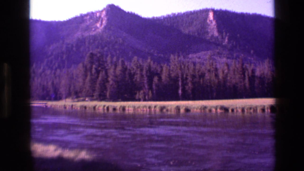 1972 Idaho home-movie frame with a distant rocky peak, forested slope, grassy bank, and purple-toned water foreground.
