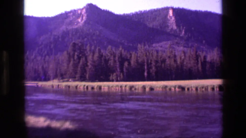 1972 Idaho home-movie frame with a distant rocky peak, forested slope, grassy bank, and purple-toned water foreground.