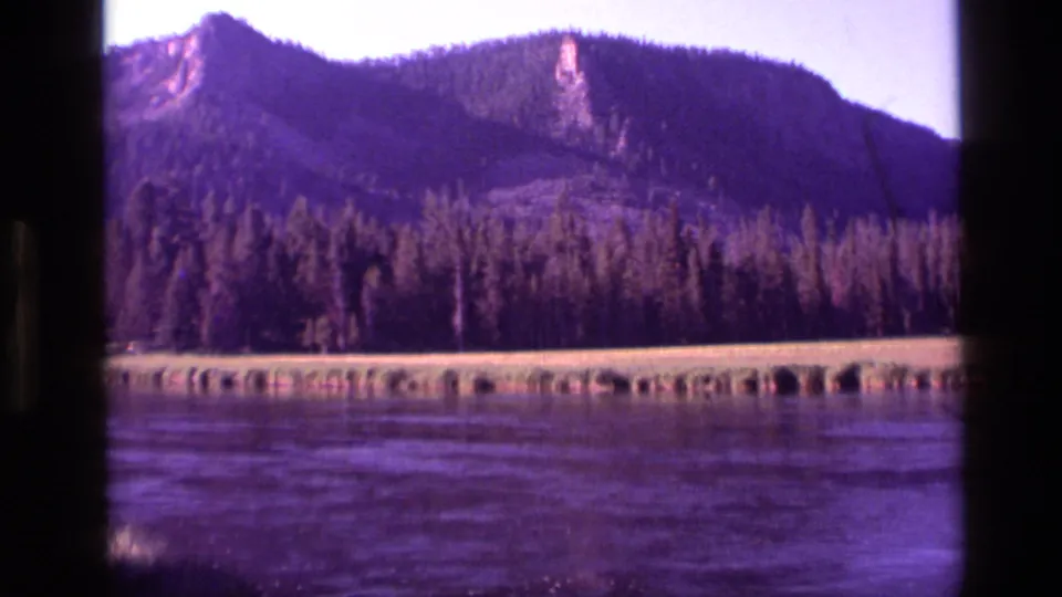1972 Idaho 8mm frame showing evergreen forest, a low grassy shore, purple-blue water, and a rugged mountain ridge in soft focus.