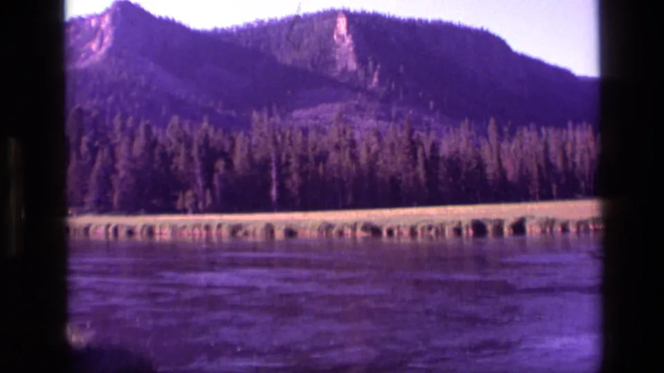1972 Idaho home-movie frame with a broad water foreground, forested shore, rocky mountain face, and visible 8mm color shift.