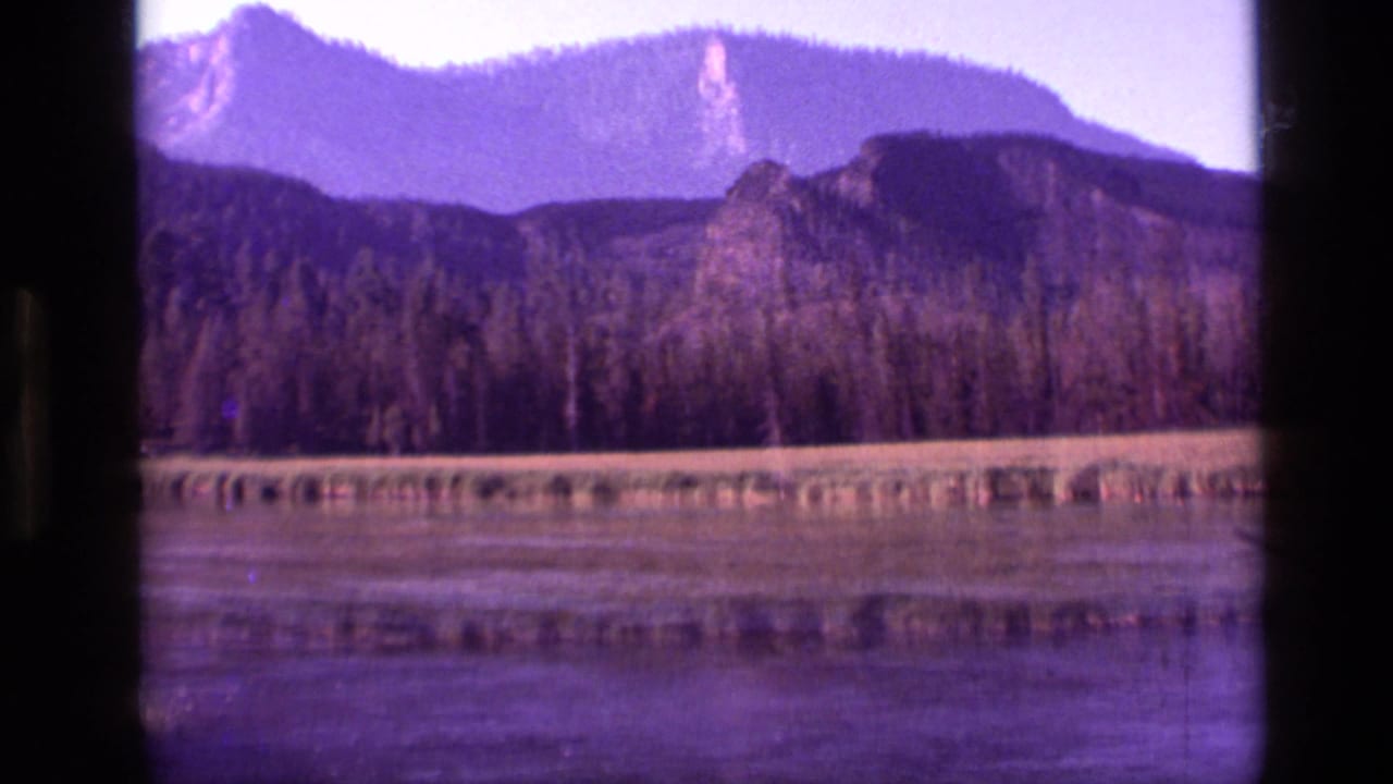 1972 Idaho 8mm frame showing calm water, meadow edge, conifer forest, and rocky mountain ridges under clear sky.