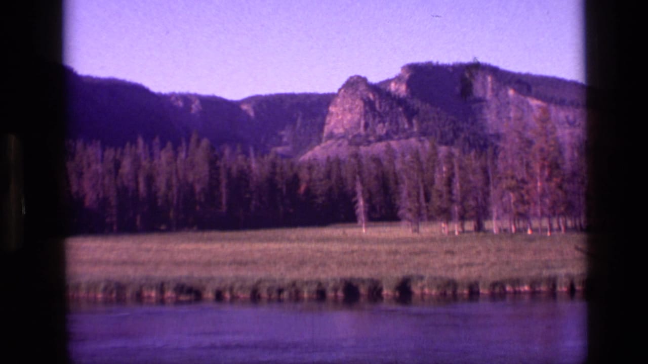 1972 Idaho home-movie view across still water toward a grassy bank, dense trees, and a rocky mountain formation.