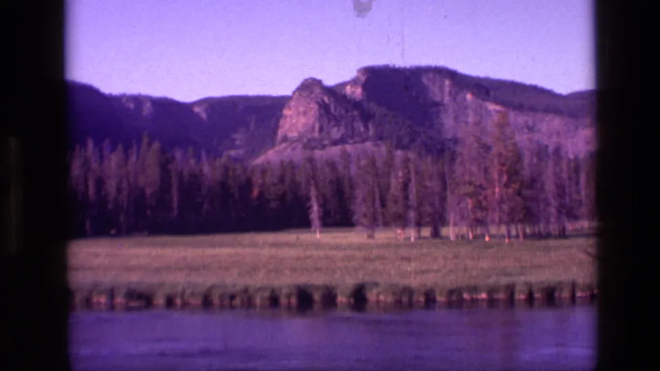 1972 Idaho 8mm landscape frame with calm river water, meadow strip, conifer forest, pale trunks, and rocky hills.