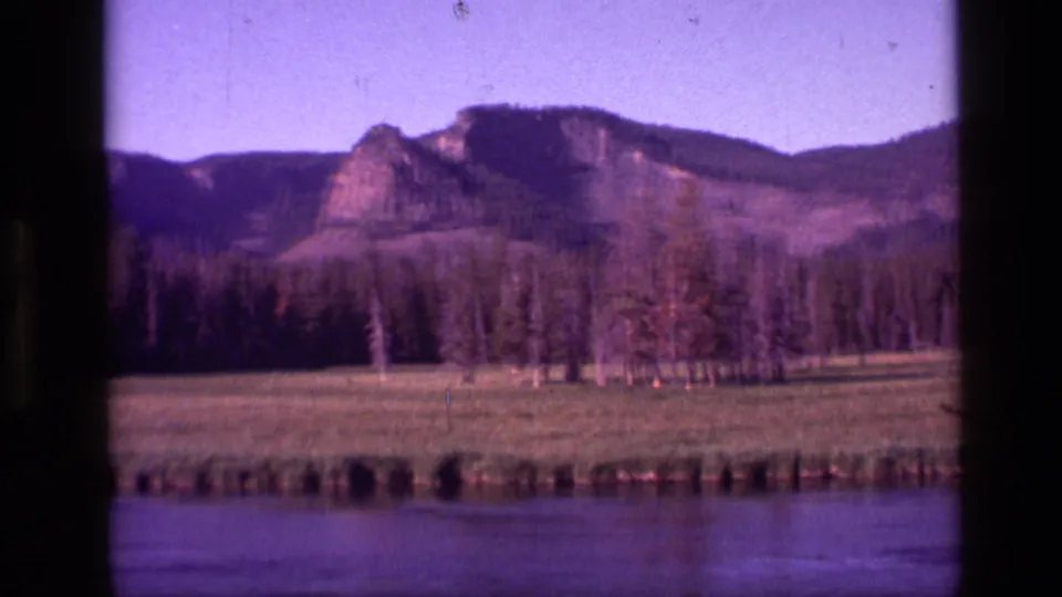 1972 Idaho home-movie frame showing a quiet river foreground with grass, trees, and a rocky mountain slope beyond.