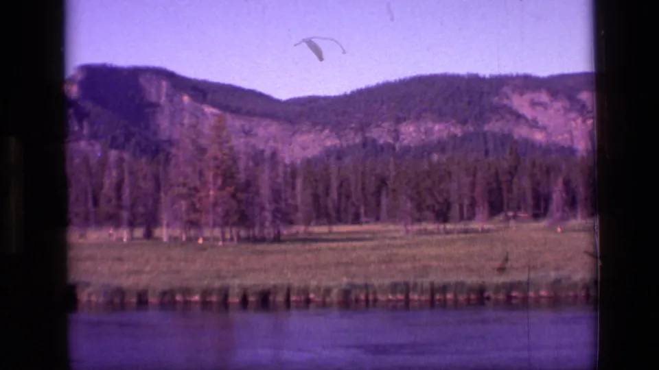 1972 Idaho 8mm frame with river water in the foreground, meadow and conifers mid-frame, and a mountain range beyond.