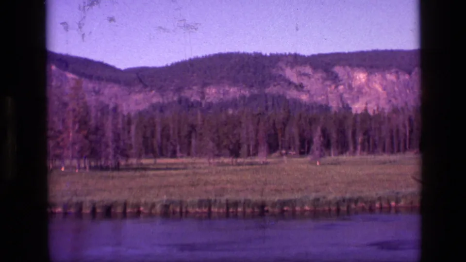 1972 Idaho 8mm frame showing a wide river foreground, grassy meadow, dense pine forest, and rocky hillside under clear sky.