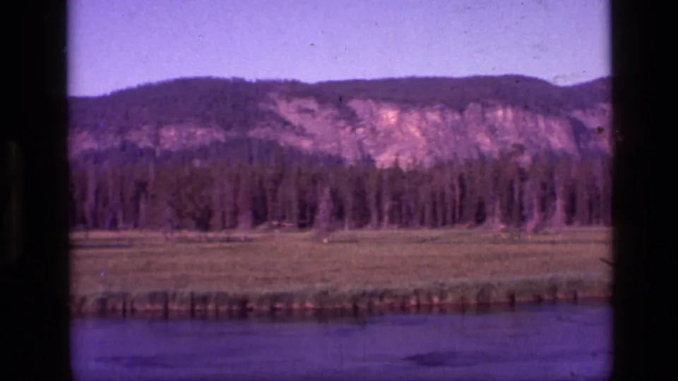 1972 Idaho 8mm frame with a river foreground, open meadow, conifer forest, and sunlit rocky mountain slope in the distance.