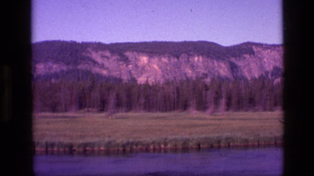 1972 Idaho 8mm frame showing water, meadow, dark forest, and a rugged hillside under a purple-tinted sky.