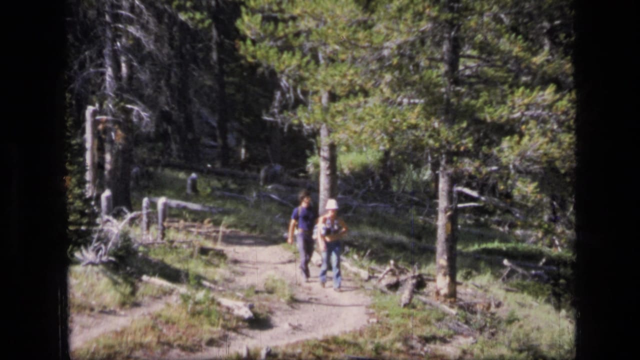 The couple continues along a Scapegoat Wilderness forest path, with the woman in a light hat and backpack visible beside the man.