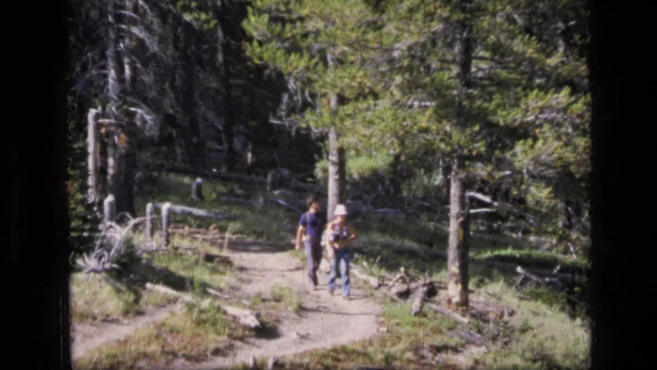 The couple continues along a Scapegoat Wilderness forest path, with the woman in a light hat and backpack visible beside the man.