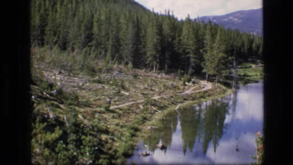 1973 Montana wilderness home-movie frame with lake reflections, pine-covered slopes, meadow clearing, and a small trail.