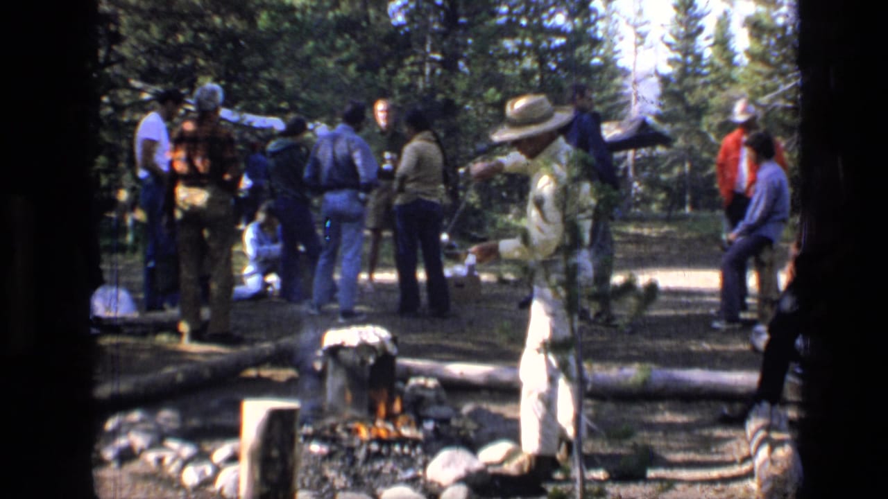 1973 8mm frame from Scapegoat Wilderness showing adults around a wooded campsite as one man tends food over a small fire.