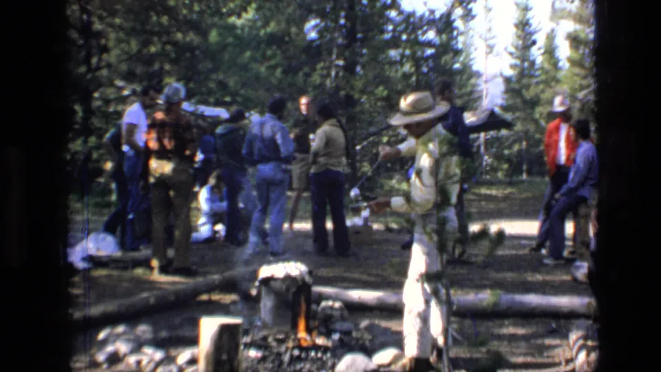 1973 Montana wilderness home-movie frame with adults in casual clothing gathered near a campfire and pine trees.