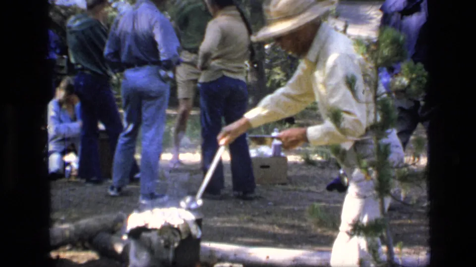 Close 1973 8mm view of a man in light clothing tending food over a campsite fire as others stand nearby.