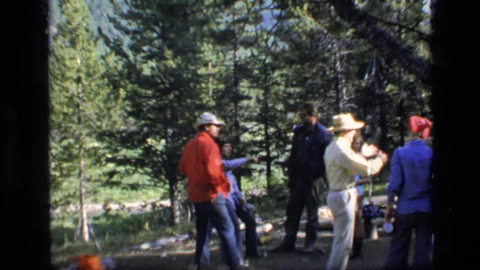 1973 Scapegoat Wilderness frame showing adults talking in a pine clearing near camp items after or around a campfire scene.