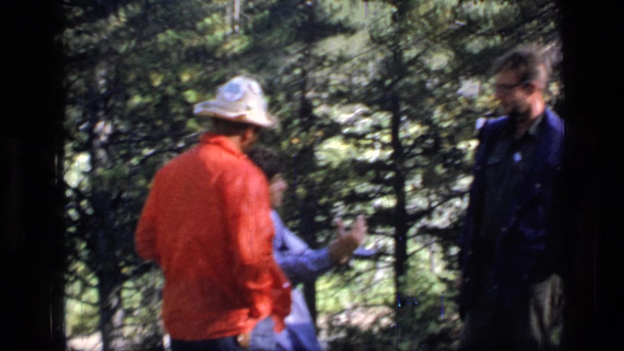 Adults in outdoor clothing talk in a wooded area, with one person gesturing while seated or crouched.