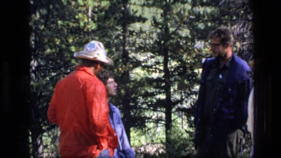 The 1973 Scapegoat Wilderness footage shows adults clustered near conifers, with one red-jacketed figure turned toward the group.
