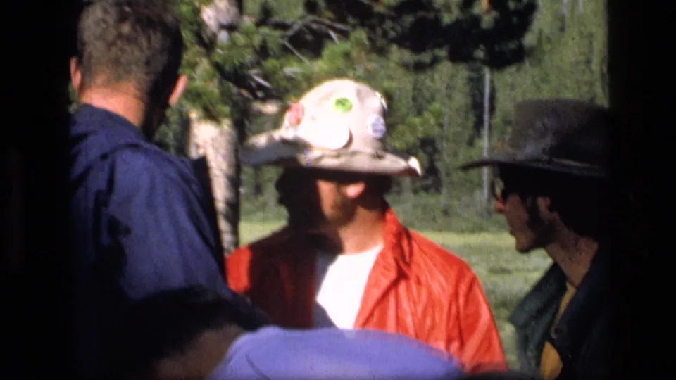 A closer 8mm frame shows adults in outdoor hats and jackets speaking together in the 1973 Montana wilderness setting.