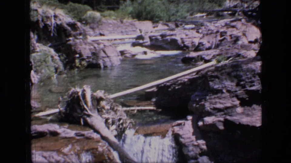 1973 8mm view of a rocky Scapegoat Wilderness stream, with a pool, cascade, fallen timber, and dark film gate edges.